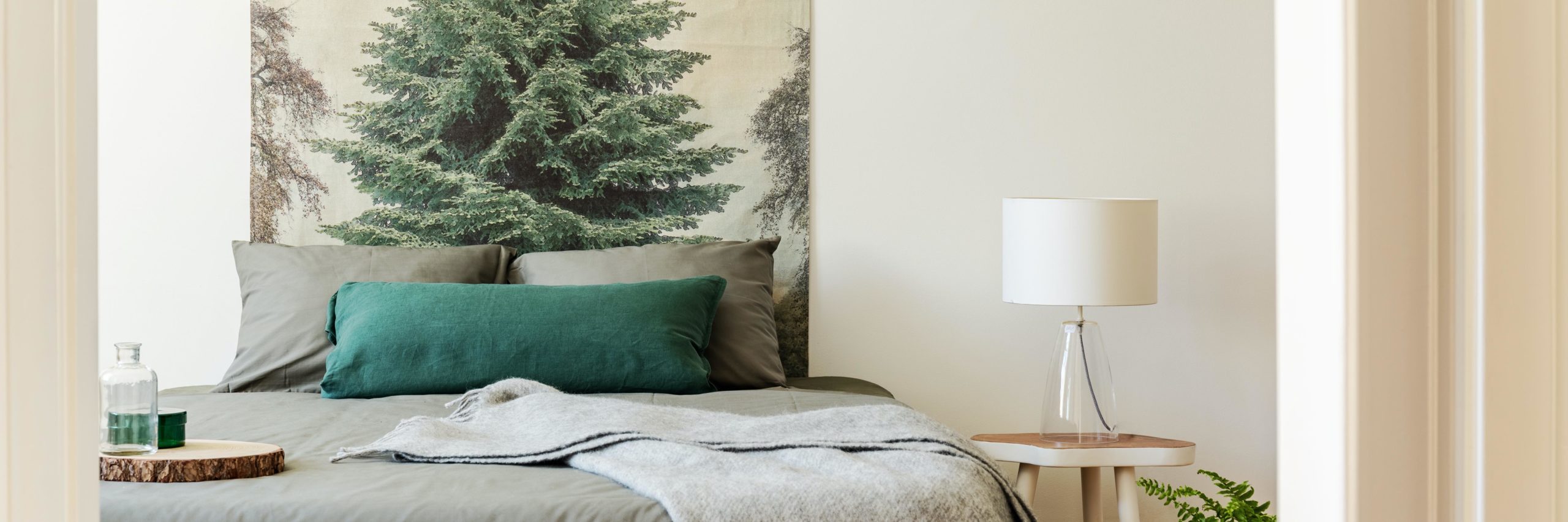 Real photo of bedside table with glass lamp standing by the double bed with grey bedclothes in bright bedroom interior with forest tree poster